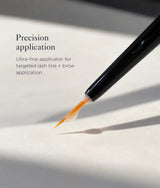 Close-up of a black precision applicator brush tip with clear gel and orange-brown pigment, shown applying product to a white surface. Text on the left reads 'Precision application' and 'Ultra-fine applicator for targeted lash line + brow application'.
