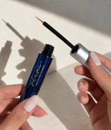 A close-up of a hand holding an open Plum Elite Prostaglandin-Free Lash & Brow Enhancing serum, person is holding the applicator wand, with a beige background and soft shadows.
