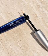 A close-up of a blue and silver Plume Hair & Lash Science lash & brow serum with its applicator wand, set diagonally on a textured beige stone surface.
